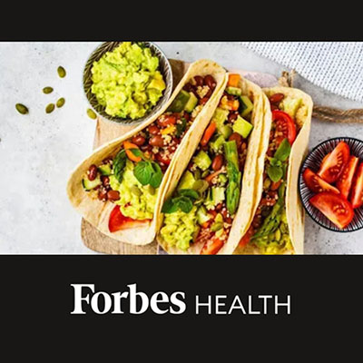Image of soft-shell tacos propped up by small bowls of guacamole and tomatoes. Forbes Health logo is underneath the image.
