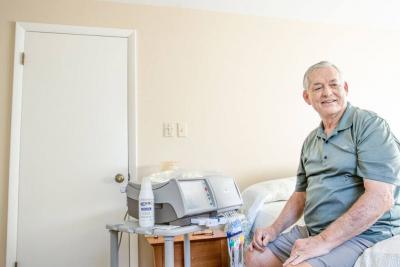 male sits on his bed at home with a home dialysis machine set up next to him
