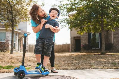 Woman holds and laughs with young boy as they play outside with a toy scooter