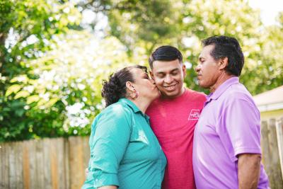 Two-time kidney transplant recipient Jose stands with his parents, he mom kisses him on the cheek. 