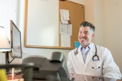 Nephrologist  wearing a white coat that has the DaVita logo sits at a desk while looking up and smiling.