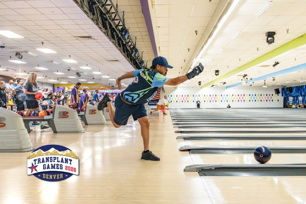 An action shot of a bowler at the Transplant Games. In the center of the photo is a single bowler as he releases a bowling ball down the lane. In the background, other participants look on and prepare to bowl.