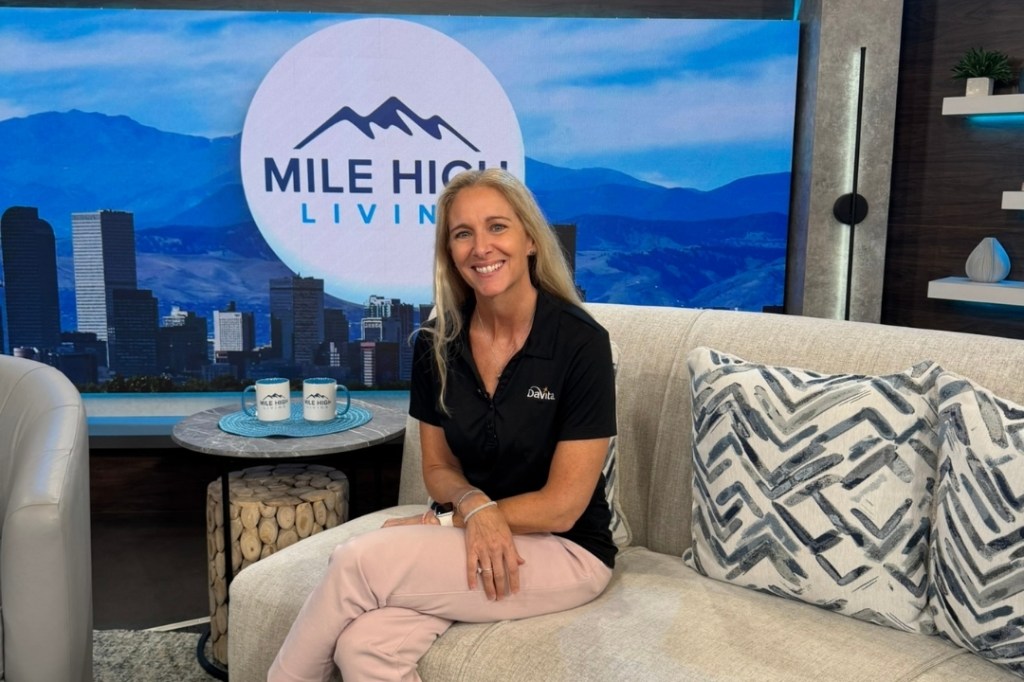 Angela Laino sits on a couch at the Mile High Living set as she smiles at the camera.