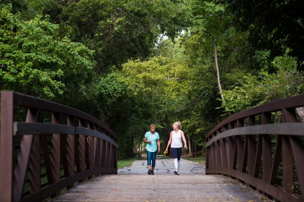 Two women walk together along a foot bridge surrounded by lush, green trees.