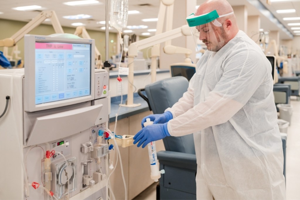 A DaVita teammate wears a clinical visor, gloves and white gown while he preps a dialysis machine with the Nipro ELISIO-HX dialyzer for middle molecule clearance.