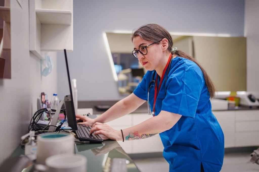 A woman in blue scrubs reviews results on a computer while standing in a medical lab setting.